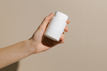 Female hand holding a white mockup jar with dietary supplement capsules on a beige background. Concept of pharmacy and medicine, health care