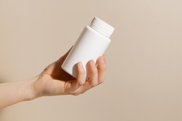 Female hand holding white plastic mockup pill jar on beige isolated background. Concept of vitamins for beauty and health, dietary supplements and pharmacy.