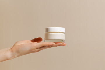 Female hand holding white glass mockup jar of face cream on beige isolated background. Concept of beauty products, skin care, nutrition and hydration