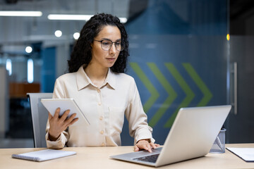 Focused Hispanic businesswoman using a laptop and holding a tablet at her modern office workspace....