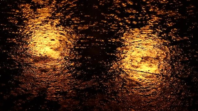 inquadratura in primo piano, di notte, della pioggia che cade intensamente sull'asfalto nero ed illuminato di arancione dal riflesso di alcuni lampioni stradali, durante un temporale