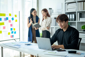 A man is working on a laptop in front of a white board with colorful notes. He is surrounded by two other people, one of whom is reading a book
