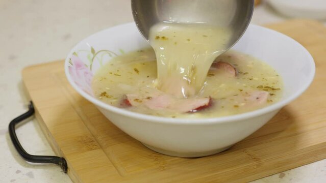 Pouring traditional Polish zurek soup or white borscht with sausages and eggs. Polish Easter soup in white bowl.