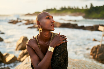Non-binary black person in luxury dress, golden jewelry on beach rocks in ocean. Trans ethnic...