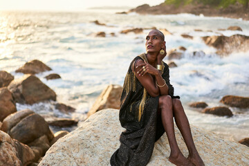 Non-binary ethnic fashion model in dress, brass jewelry sits on rocks by ocean. Trans sexual black...