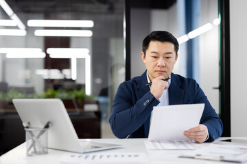 Focused Asian businessman with glasses examining paperwork at his desk in a contemporary office...