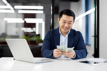 Smiling Asian businessman sitting at a modern office desk, counting US dollar bills. Concept of...