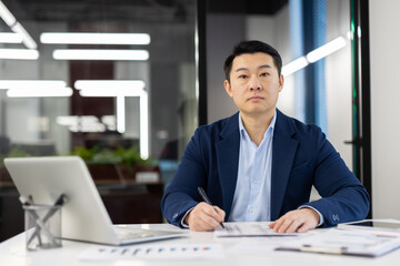 Focused Asian businessman in a well-fitted blue suit working at his office desk surrounded by...