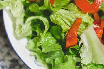 vista macro da sopra e da vicino di varie foglie verdi d'insalata insieme a dei pezzi di peperone rosso, in un piatto bianco, su un tavolo di marmo grigio