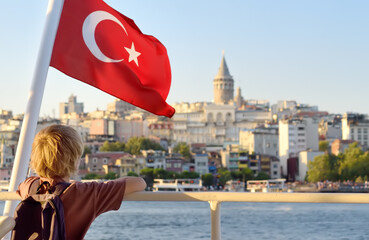 Preteen tourist boy is admiring of view of Golden Horn seafront from tour boat at sunset. Far away is famous Galata Tower. Busy urban cityscape of Istanbul with turkish flag on foreground