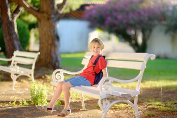 Cute blonde preteen tourist boy is sitting on a white bench in beautiful old park at summer sunset. Resort for family vacation.
