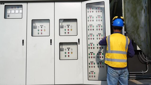 Asian male engineer wearing a reflective jacket, hard hat, walking work electrical cabinet, inspecting electrical systems, building on rooftop of construction building.
