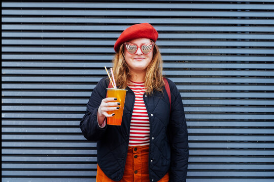 Smiling plus size woman in heart shaped sunglasses and bright clothes drinking sugar flavored tapioca bubble tea near striped urban wall. Happy fashionable hipster overweight girl. Street fashion.