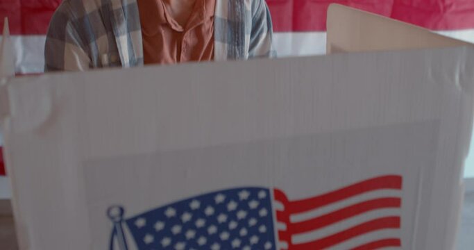 Asian man in a voting booth at a polling station with an American flag in the background