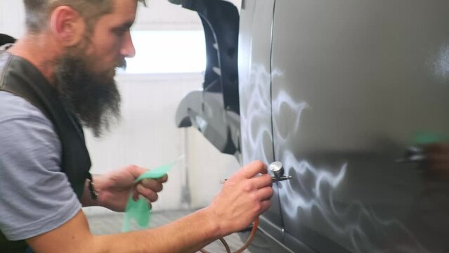 Bearded, white-haired, middle-aged master airbrushing car body door side with smooth curves using silicone round shape sample. Drawing wavy white lines of air flows on black background of automobile.