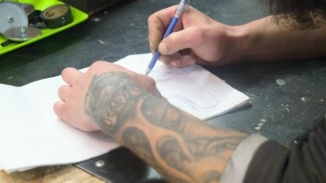 Process of drawing motorcycle helmet sketch. Tattooed bearded biker hands with blue ballpoint pen making sketch in notebook on black stone table in motorcyclist auto repair shop. Close up side view.