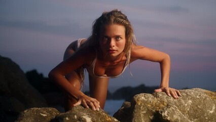 Woman in swimsuit crawls on rocky shore, channels wild instinct, primal strength in nature. Intense gaze, motion blur, sunset light captures raw feminity, survival theme, beach landscape backdrop.