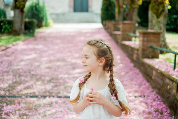 Happy pretty girl 6 years old in white dress near blooming pink sakura. The child is walking in the park. Spring.