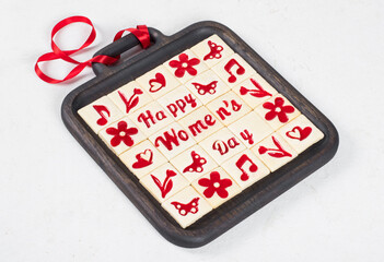 Women's Day cookies. Little square shortbread cookies with marmalade filling of a thematic shape: Happy Women's Day, flower, heart, butterfly, notes. On a wooden plate with a red satin ribbon. 