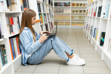 Schoolgirl sitting on a floor among books and using a laptop in bookstore or library. School education.