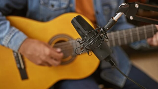 A male musician strums an acoustic guitar during a recording session in a studio, with microphone in focus.