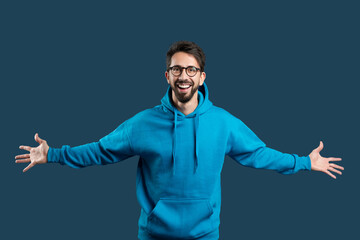 A man wearing a blue hoodie stands with his hands extended outward. He appears to be gesturing or reaching for something, with a focused expression on his face.