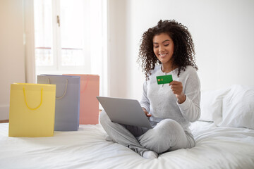 Hispanic woman is seated on a bed, holding a credit card in one hand and a laptop in the other. She...