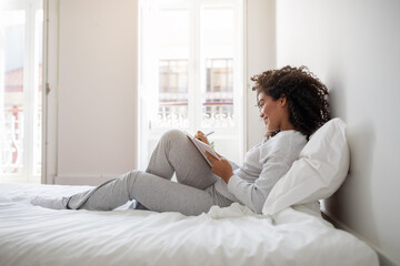 Hispanic woman is seated on a bed, focused on writing in a notebook. She appears to be deep in thought, jotting down ideas or reflections. The room is simple, with neutral decor and soft lighting.