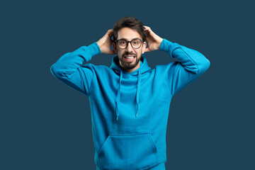 A man wearing a blue hoodie is standing with his hands pressed against his ears, appearing to be in...