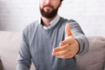 A man with a beard is seen reaching out his hand to offer a handshake, wearing a casual gray shirt and is situated indoors, with a light-colored couch partially visible behind him, cropped