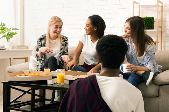 A small group of multiracial friends is seated around a coffee table in a well-lit living room, sharing laughter and stories over a meal of pizza and refreshing beverages.