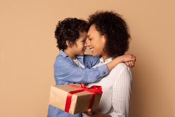 Cute African American little boy greeting his mom with Mothers Day, giving her wrapped box and...