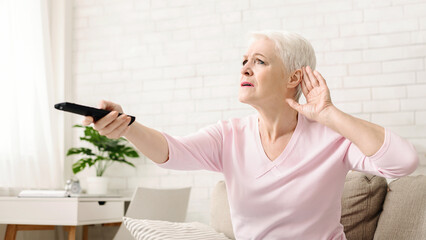 Senior woman cupping her hand behind ear to hear better to the television, making sound louder,...