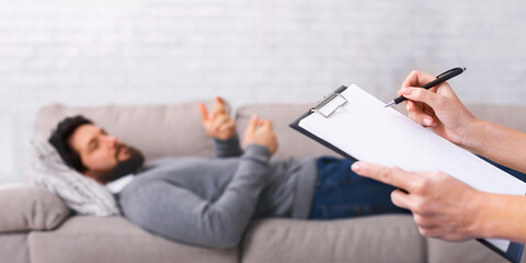 A young man lies comfortably on a gray couch, gesturing with his hands while speaking to someone...