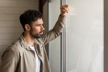 A thoughtful young man gazes out of a clear glass window, his hand gently resting on the pane, with...