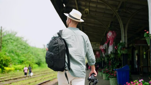 Asian travel blogger explores Haputale railway station with backpack, camera in hand. Man observes trains, local culture in Sri Lanka setting. Content creator documents authentic travel experience.