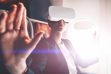 Young woman in suit wearing virtual reality headset in airplane and gesturing with hands to control...