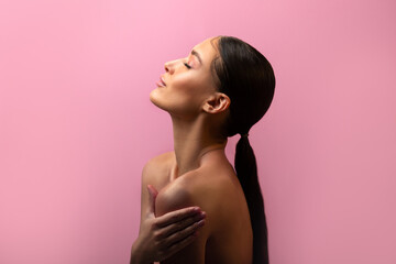 Charming lady with ponytail hug her naked shoulders, posing with closed eyes isolated on pink studio background with copy space, side view