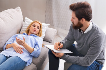A young woman lies on a couch, looking pensive and speaking, while man therapist with a beard attentively listens and takes notes on a clipboard.