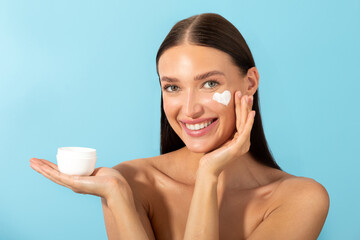 Skin hydration ad. Cheerful smiling lady holding jar and applying moisturizing cream on face over blue studio background