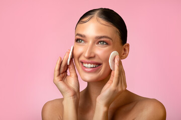 Pretty young lady touching her face with cotton pads, removing makeup with cleansing milk, using...