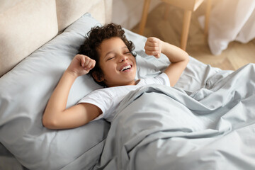 African American young boy is lying comfortably in his bed, with a soft smile on his face. He looks content and relaxed, enjoying a moment of peace before sleep, stretching