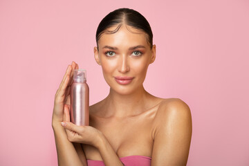 Beautiful European woman with stylish hairstyle holding hair spray bottle, posing on pink studio background and looking at camera