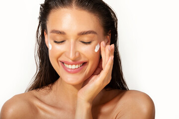 Skin care concept. Happy European woman applying cosmetic moisturizing cream on her face and smiling with closed eyes on white background