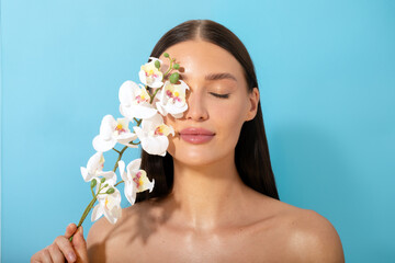 Face skin care and make up. Young lady with nude makeup and perfect glowing skin, posing holding orchid flowers, blue background