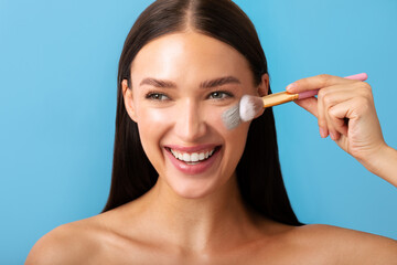 Happy woman with brush for makeup, applying face mask for skincare, standing on blue background. Cosmetics product and healthy skin motivation