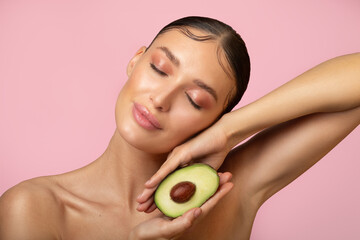 Attractive lady posing with avocado half in hands, enjoying organic ingredients for cosmetics, standing on pink studio background