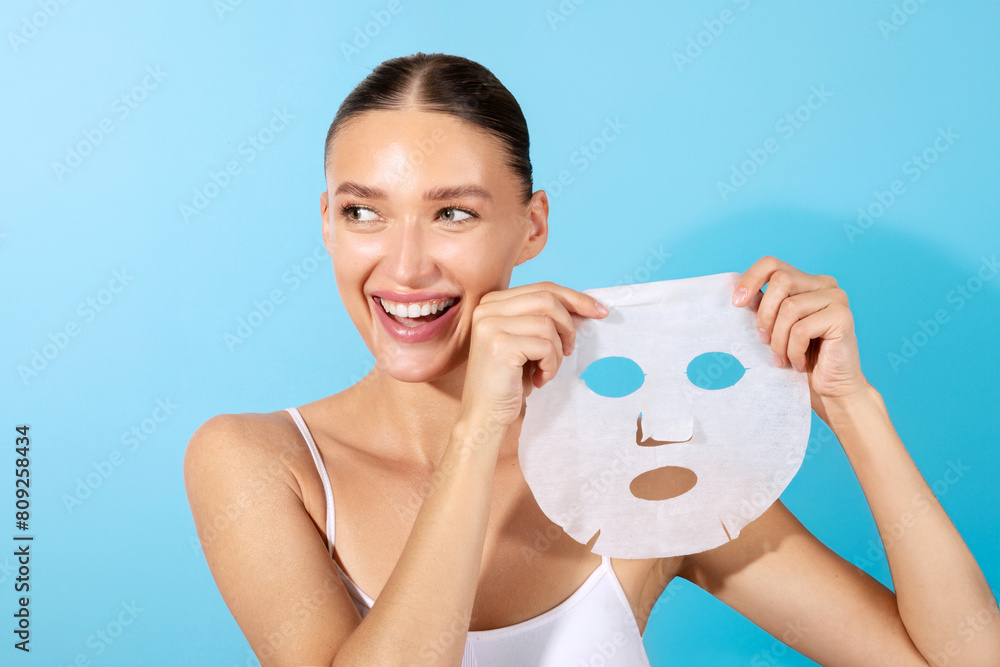 Wall mural Attractive European woman holding paper sheet mask, enjoys beauty skin care, smiling positively, standing isolated on blue background