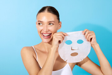Attractive European woman holding paper sheet mask, enjoys beauty skin care, smiling positively, standing isolated on blue background