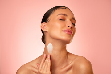 Sensual young woman with applying body cream or lotion on her neck, standing against pink studio...